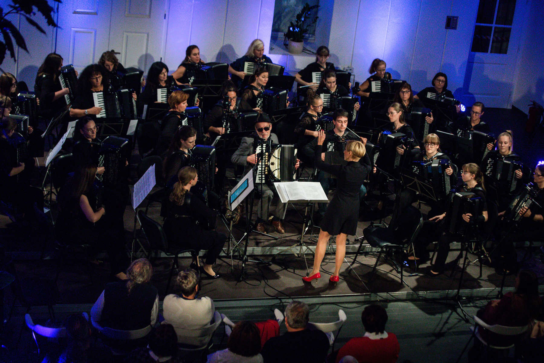 Eine Gruppe von 30 Frauen spielt Akkordeon, alle konzentriert und harmonisch zusammen. In der Mitte steht die Dirigentin, die mit ausdrucksvollen Gesten die Musikerinnen führt. Die Szene strahlt Leidenschaft, Präzision und musikalische Einheit aus.