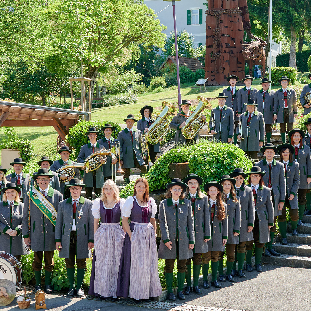 Eine große Gruppe von Menschen in traditioneller Kleidung posiert für ein Foto im Freien. Viele tragen graue Jacken, grüne Hosen und Hüte, während einige Frauen Dirndl tragen. Einige Personen halten Blechblasinstrumente. Sie stehen auf einer Treppe und einer Rasenfläche mit Bäumen und einem Gebäude im Hintergrund.