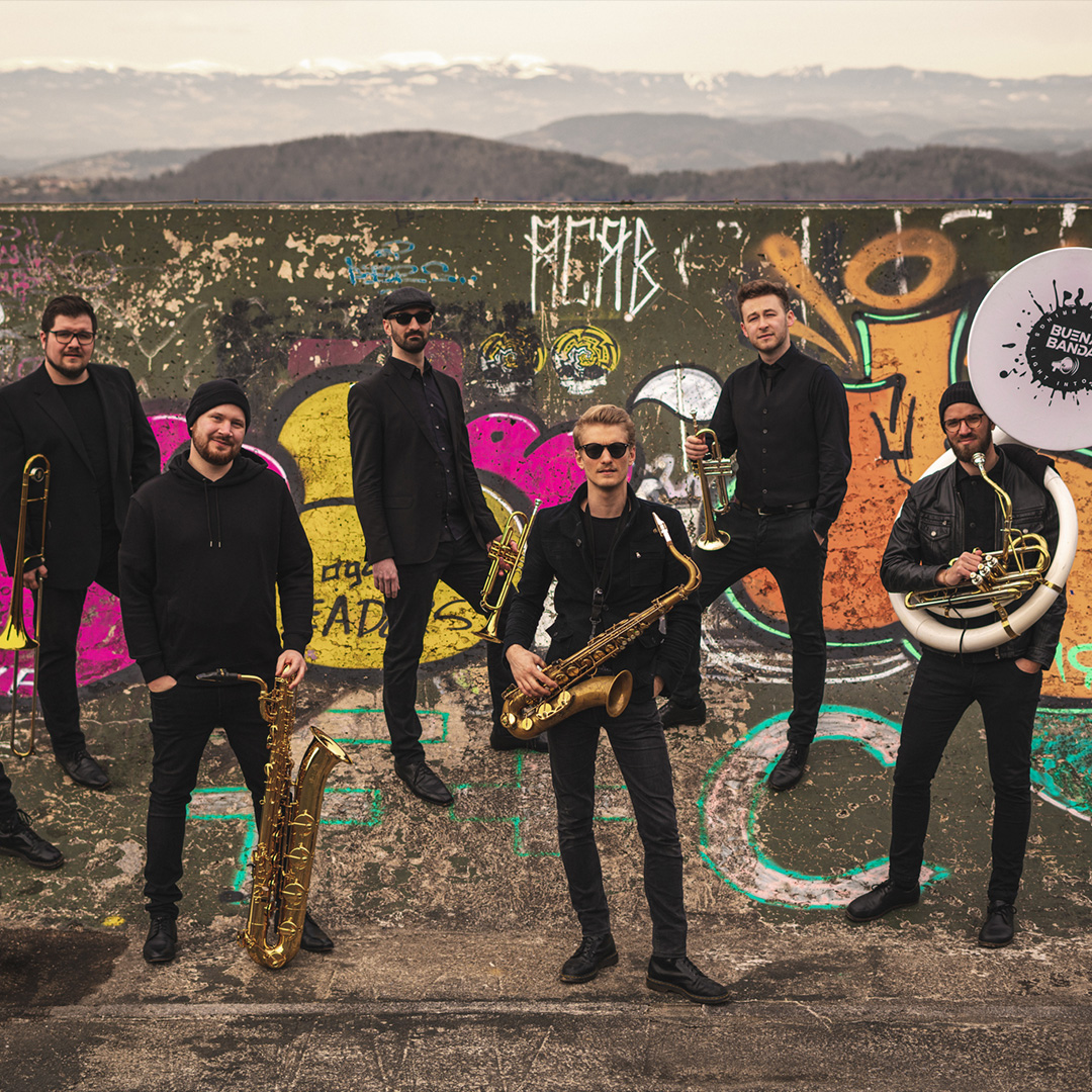 Ein Gruppenfoto der Brass-Band „Buena Banda“. Sechs Musiker in schwarzer Kleidung posieren vor einer mit bunten Graffitis besprühten Mauer. Sie halten ihre Instrumente, darunter Saxophone, Trompeten, eine Posaune und ein Sousaphon mit dem Band-Logo. Im Hintergrund ist eine weite, hügelige Landschaft unter einem bewölkten Himmel zu sehen.