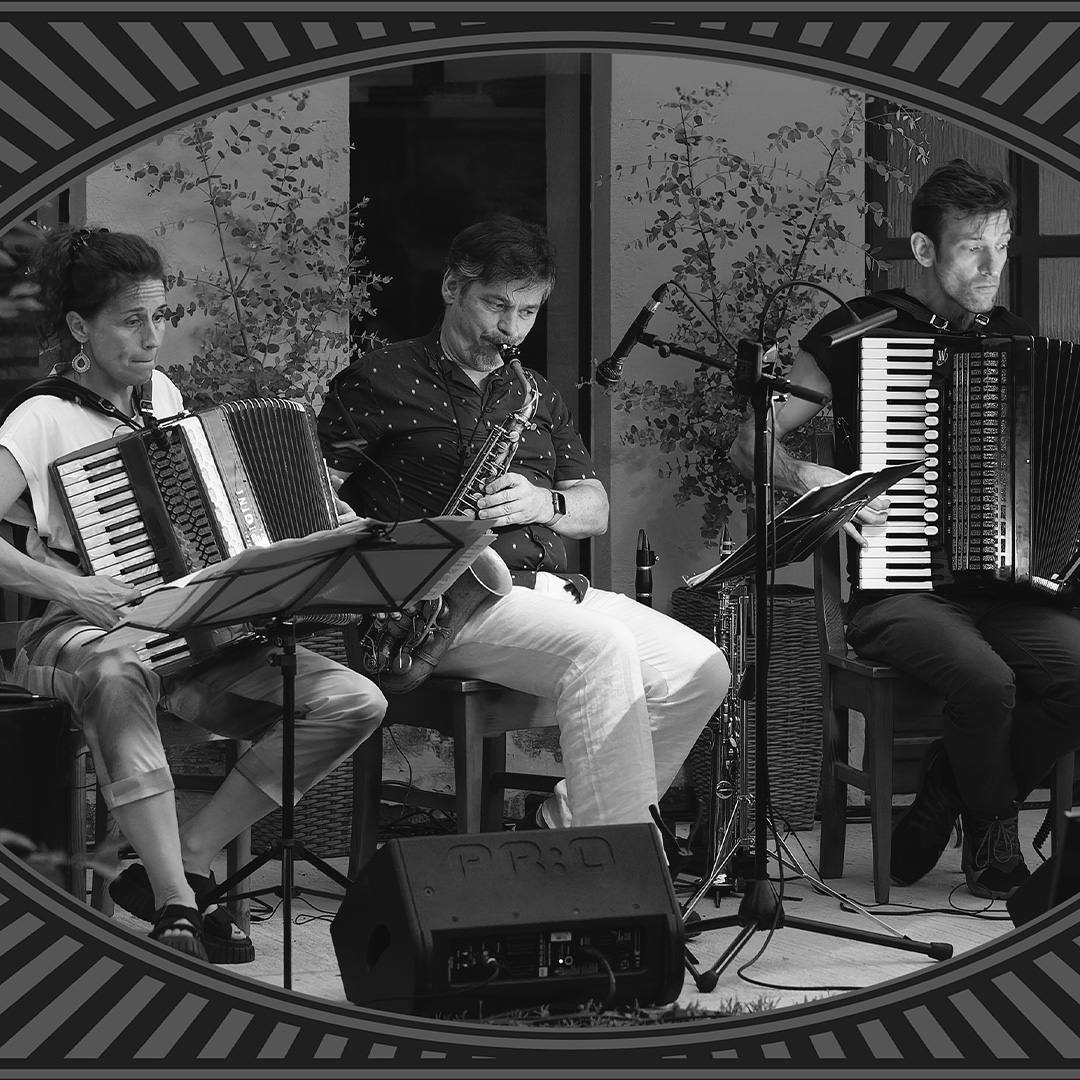 Ein Schwarz-Weiß-Foto eingerahmt in ein klassisches Stummfilm-Design. Es zeigt das Trio Franziska Hatz, Richie Winkler und Tino Klissenbauer bei einer Live-Performance. Links sitzt Franziska Hatz mit einem Akkordeon, in der Mitte Richie Winkler mit einem Saxophon und rechts Tino Klissenbauer, ebenfalls am Akkordeon. Vor ihnen stehen Notenständer und ein Bühnenmonitor. Oben rechts sind die Logos von „derFlor Fotografie“ und „Straden aktiv“ zu sehen.