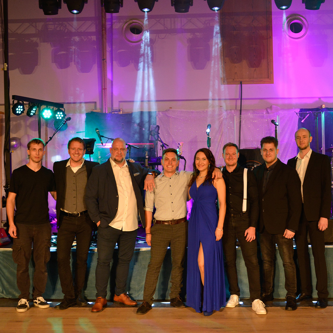 Ein Gruppenfoto der Coverband „Smash Time“ auf einer Konzertbühne. Acht Personen posieren lächelnd vor ihrem Musik-Equipment, darunter sieben Männer in legerer und eleganter schwarzer Kleidung sowie eine Sängerin in einem langen, leuchtend blauen Kleid in der Mitte. Die Bühne ist professionell mit violettem und grünem Showlicht beleuchtet, im Hintergrund sind Schlagzeug, Verstärker und Lichtanlagen zu sehen.