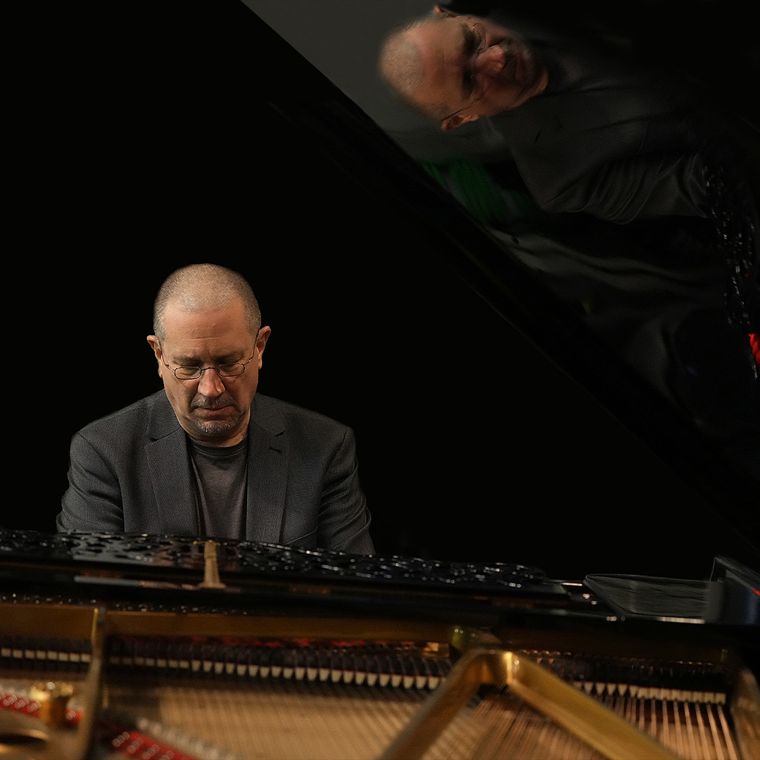 Ein Porträtfoto des Pianisten Chris Jarrett beim Klavierspielen vor schwarzem Hintergrund. Man sieht den Musiker konzentriert mit Blick nach unten auf die Tastatur, während sich sein Gesicht und Oberkörper im hochgeklappten, glänzenden Deckel des Flügels spiegeln. Im Vordergrund ist ein Teil des Innenlebens des Klaviers mit den Saiten zu sehen.