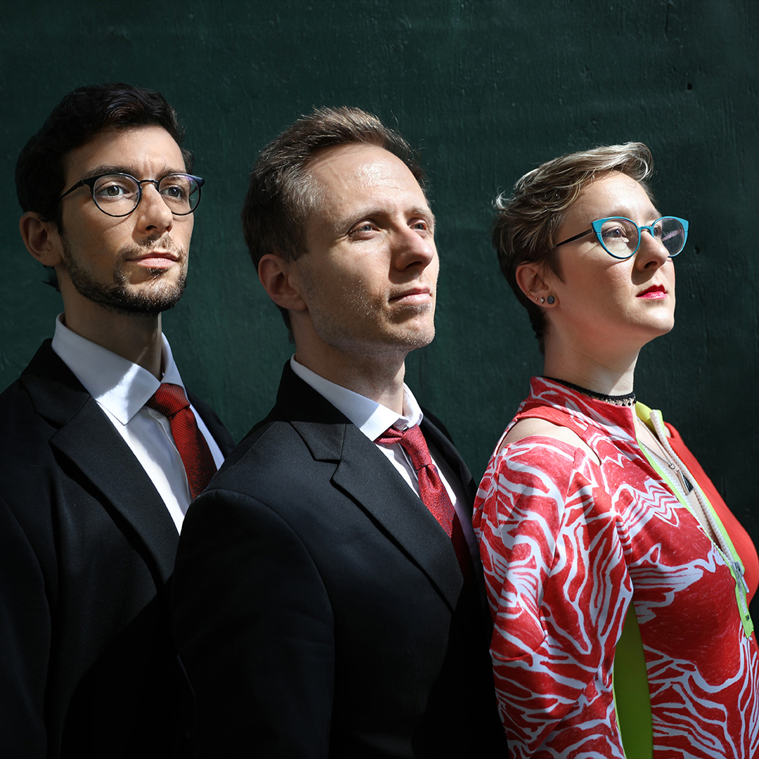 Promocijska fotografija skupine Jean John Trio feat. Kelly Green & Luca Soul: trije glasbeniki stojijo drug ob drugem v profilu in gledajo v isto smer. Dva moška v elegantnih temnih oblekah z rdečima kravatama ter pevka in pianistka s kratkimi lasmi in očali v rdeče-beli vzorčasti obleki. Dramatična osvetlitev poudarja resnost in umetniški značaj jazz koncerta v Zehnerhaus Bad Radkersburg.