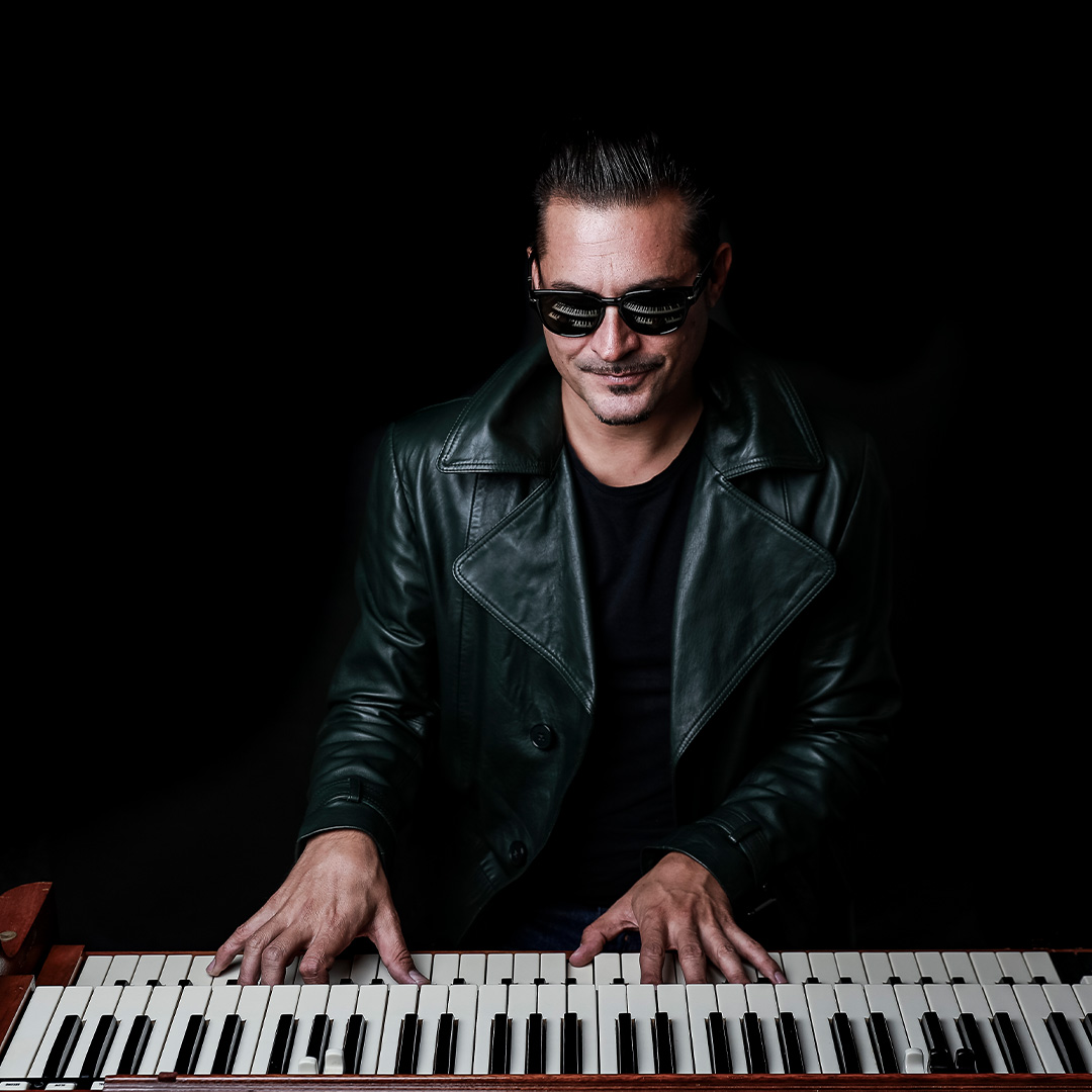 Ein Mann mit Sonnenbrille und einer dunkelgrünen Lederjacke blickt lächelnd nach unten, während er mit beiden Händen auf einem Tasteninstrument spielt. Der Hintergrund ist komplett schwarz.