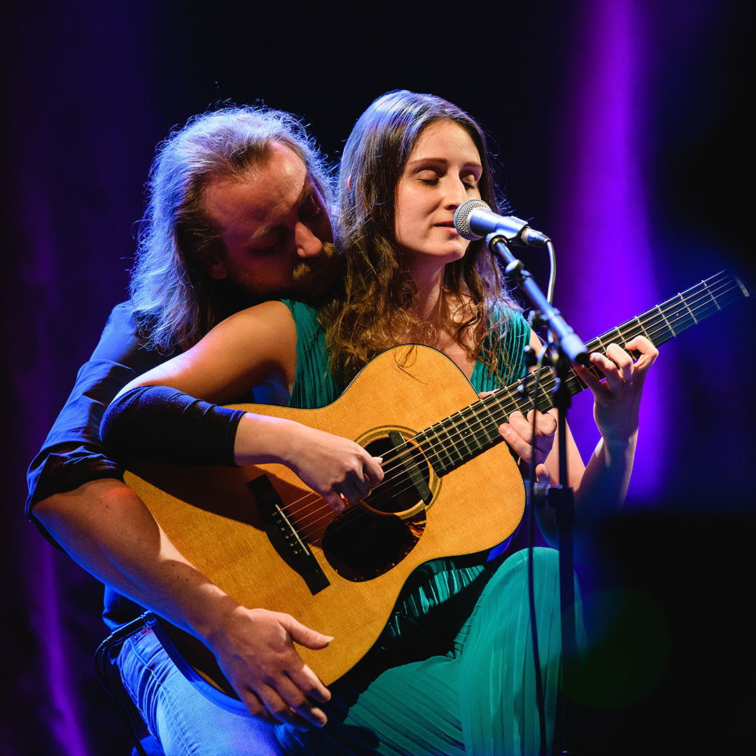 Ein Mann und eine Frau spielen gemeinsam auf einer akustischen Gitarre während eines Konzerts. Die Frau im türkisfarbenen Kleid sitzt vorne, hat die Augen geschlossen und singt in ein Mikrofon, während sie die Akkorde greift. Der Mann sitzt eng hinter ihr und zupft die Saiten. Die Bühne ist in stimmungsvolles violettes Licht getaucht.